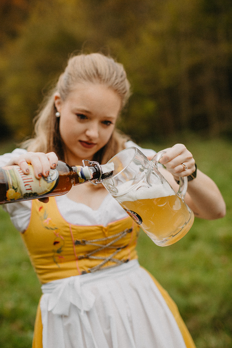 A beautiful young blonde woman pours a beer in a field near Burg Eltz wearing a traditional dirndl for these Eltz Castle couples photos in Germany