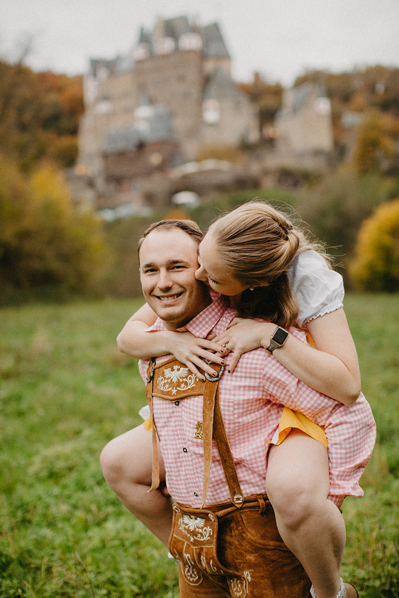 A couple embrace as he gives her a piggyback ride in a field near Burg Eltz wearing a traditional dirndl and lederhosen for these Eltz Castle couples photos in Germany