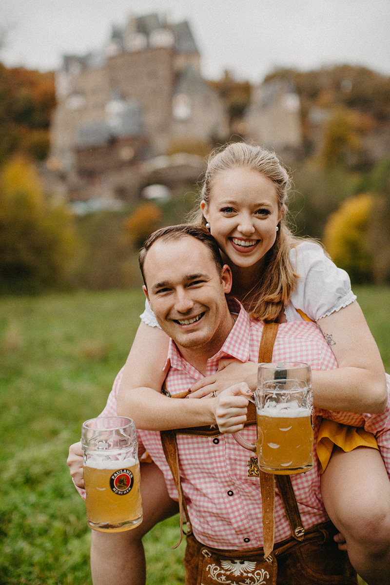 A couple embrace as he gives her a piggyback ride with beers in a field near Burg Eltz wearing a traditional dirndl and lederhosen for these Eltz Castle couples photos in Germany