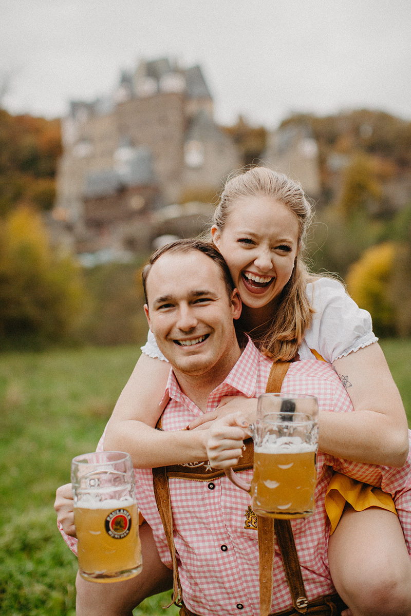 A couple embrace as he gives her a piggyback ride with beers in a field near Burg Eltz wearing a traditional dirndl and lederhosen for these Eltz Castle couples photos in Germany