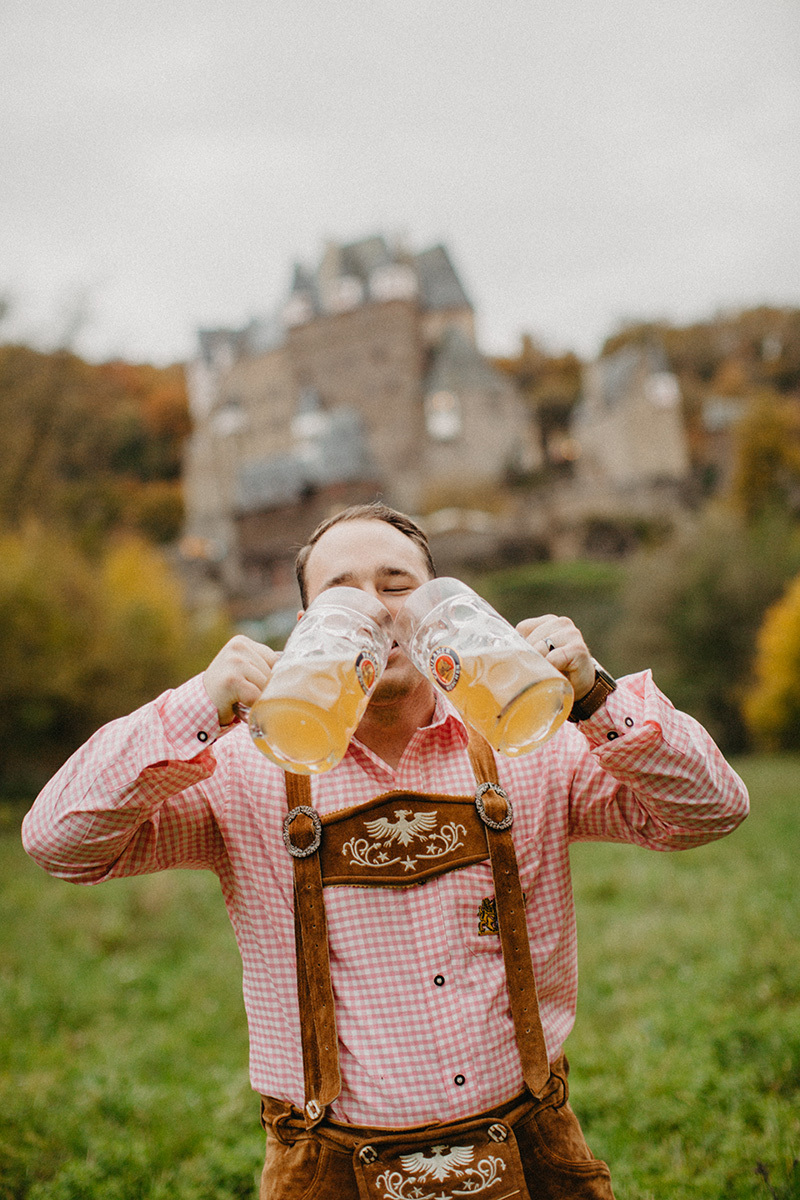A handsome young man drinks beer in a field near Burg Eltz wearing traditional lederhosen for these Eltz Castle couples photos in Germany