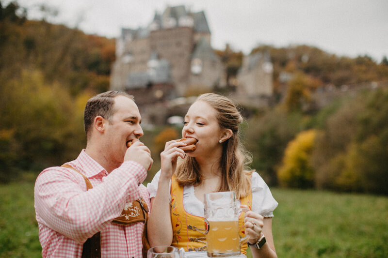 A couple eat pretzels together with mugs of beer in a field near Burg Eltz wearing a traditional dirndl and lederhosen for these Eltz Castle couples photos in Germany