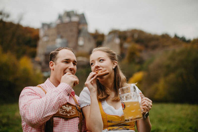 A couple eat pretzels together with mugs of beer in a field near Burg Eltz wearing a traditional dirndl and lederhosen for these Eltz Castle couples photos in Germany