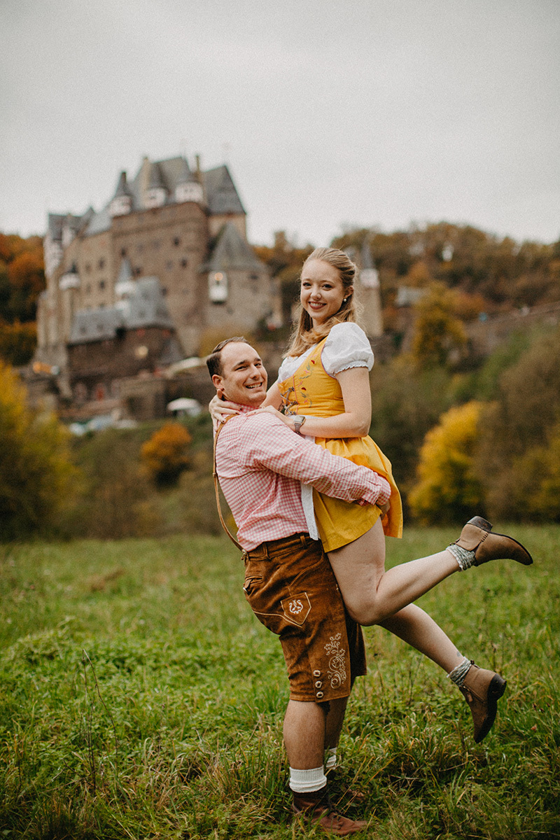 A couple embraces as he lifts her up in a field near Burg Eltz wearing a traditional dirndl and lederhosen for these Eltz Castle couples photos in Germany