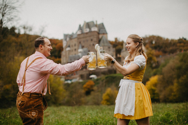 A couple cheers with mugs in a field near Burg Eltz wearing a traditional dirndl and lederhosen for these Eltz Castle couples photos in Germany