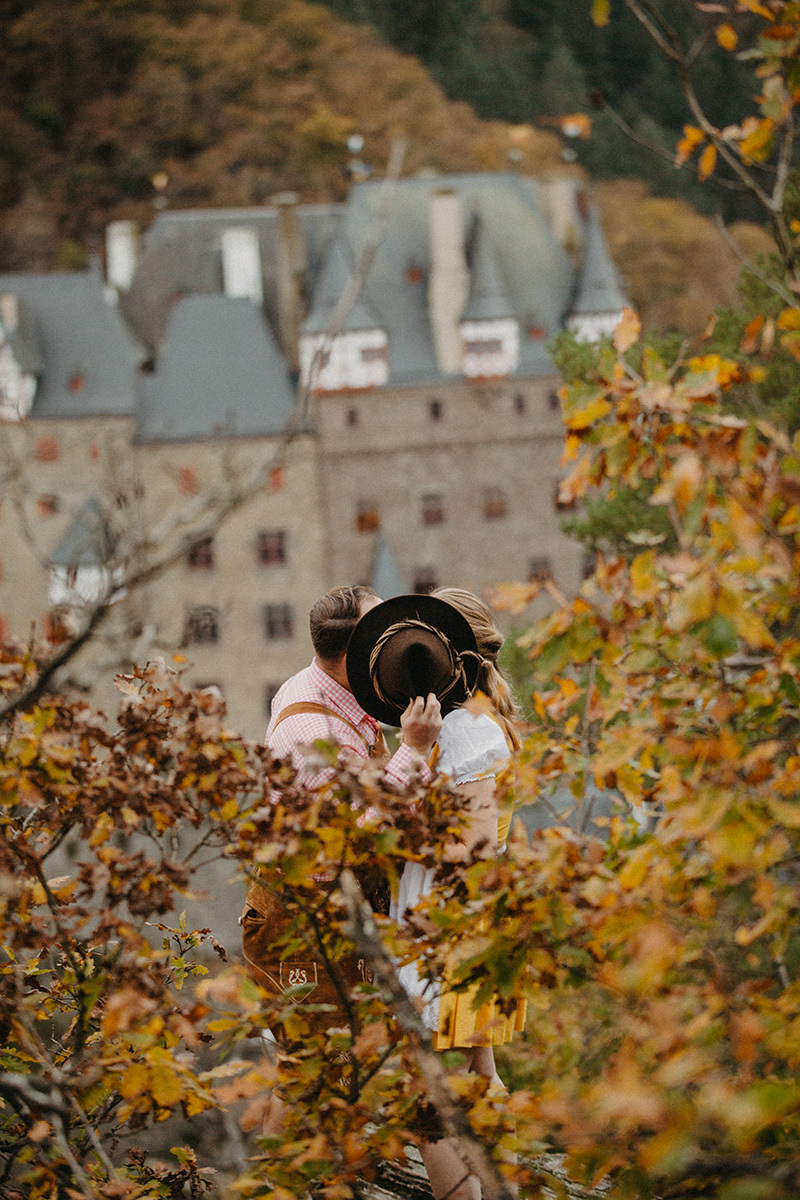 A couple embrace and kiss behind a hat near Burg Eltz wearing a traditional dirndl and lederhosen for these Eltz Castle couples photos in Germany