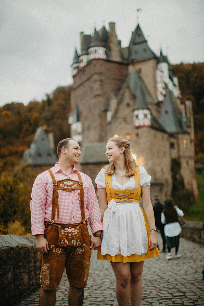 A couple holds hands in front of Burg Eltz wearing a traditional dirndl and lederhosen for these Eltz Castle couples photos in Germany