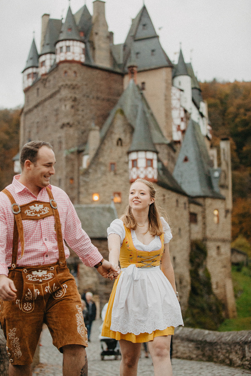 A couple holds hands in front of Burg Eltz wearing a traditional dirndl and lederhosen for these Eltz Castle couples photos in Germany