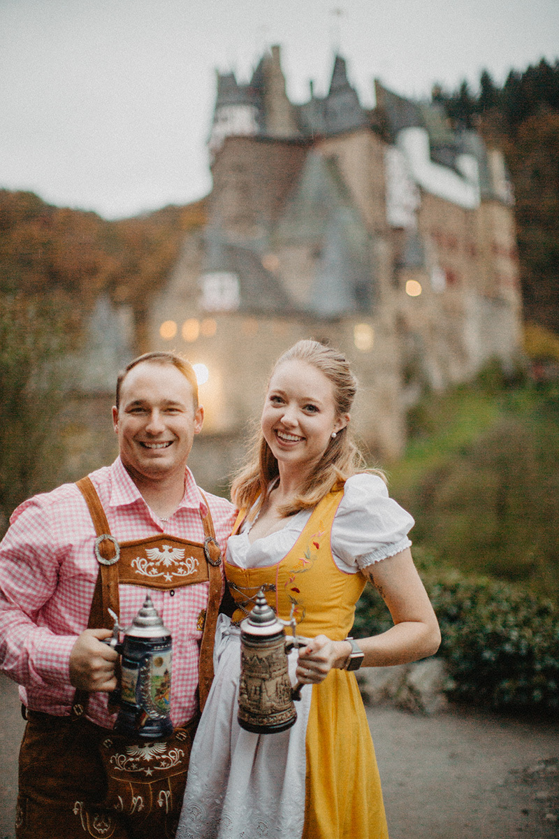 A couple embraces holding steins in front of Burg Eltz wearing a traditional dirndl and lederhosen for these Eltz Castle couples photos in Germany
