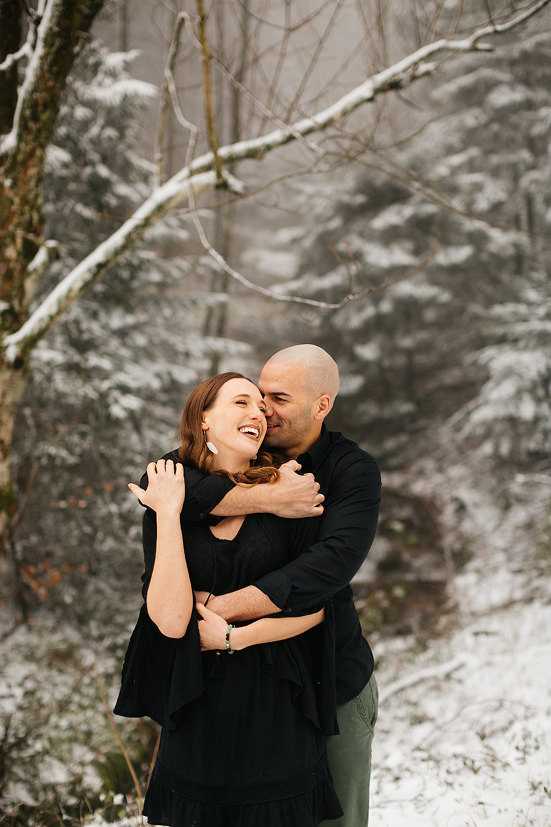 A couple embrace and laugh in a snow covered forest in Germany wearing a beautiful black dress and black button up shirt for a Black Forest engagement photography session