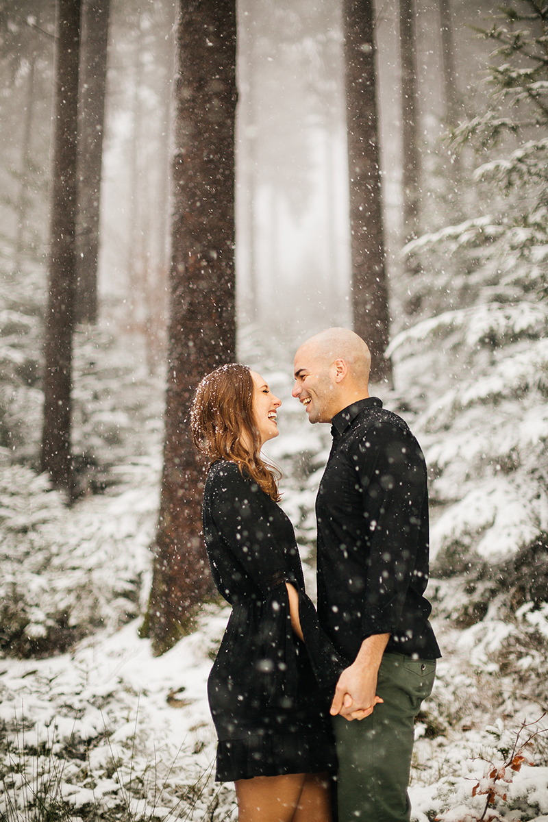 A couple hold hands in a snow covered forest in Germany wearing a beautiful black dress and black button up shirt for a Black Forest engagement photography session
