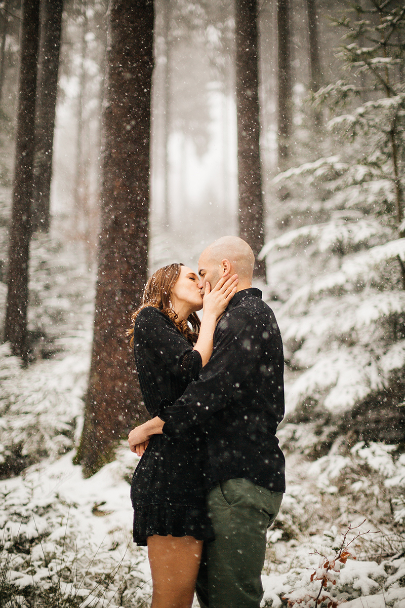 A couple embrace and kiss in a snow covered forest in Germany wearing a beautiful black dress and black button up shirt for a Black Forest engagement photography session