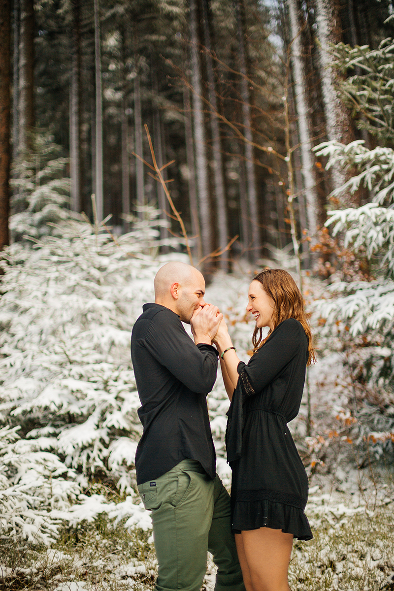 A couple embrace as he warms her handds in a snow covered forest in Germany wearing a beautiful black dress and black button up shirt for a Black Forest engagement photography session