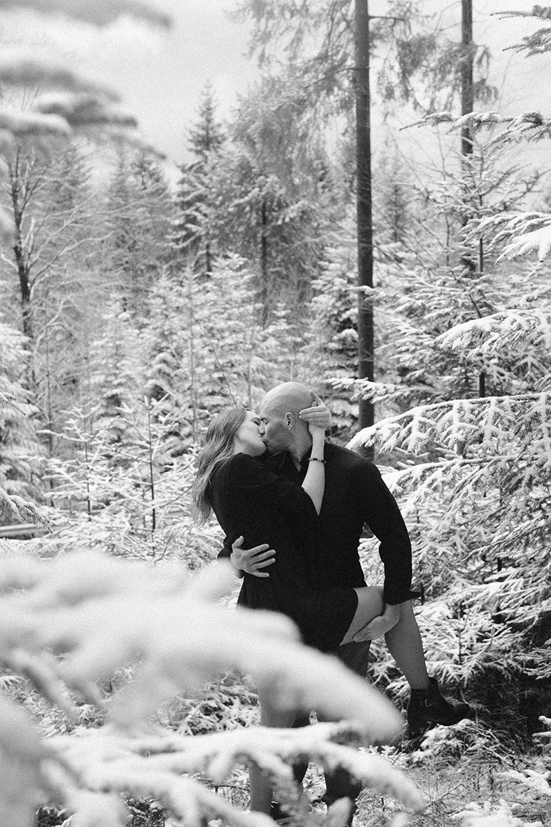 A couple embrace and kiss in a snow covered forest in Germany wearing a beautiful black dress and black button up shirt for a Black Forest engagement photography session