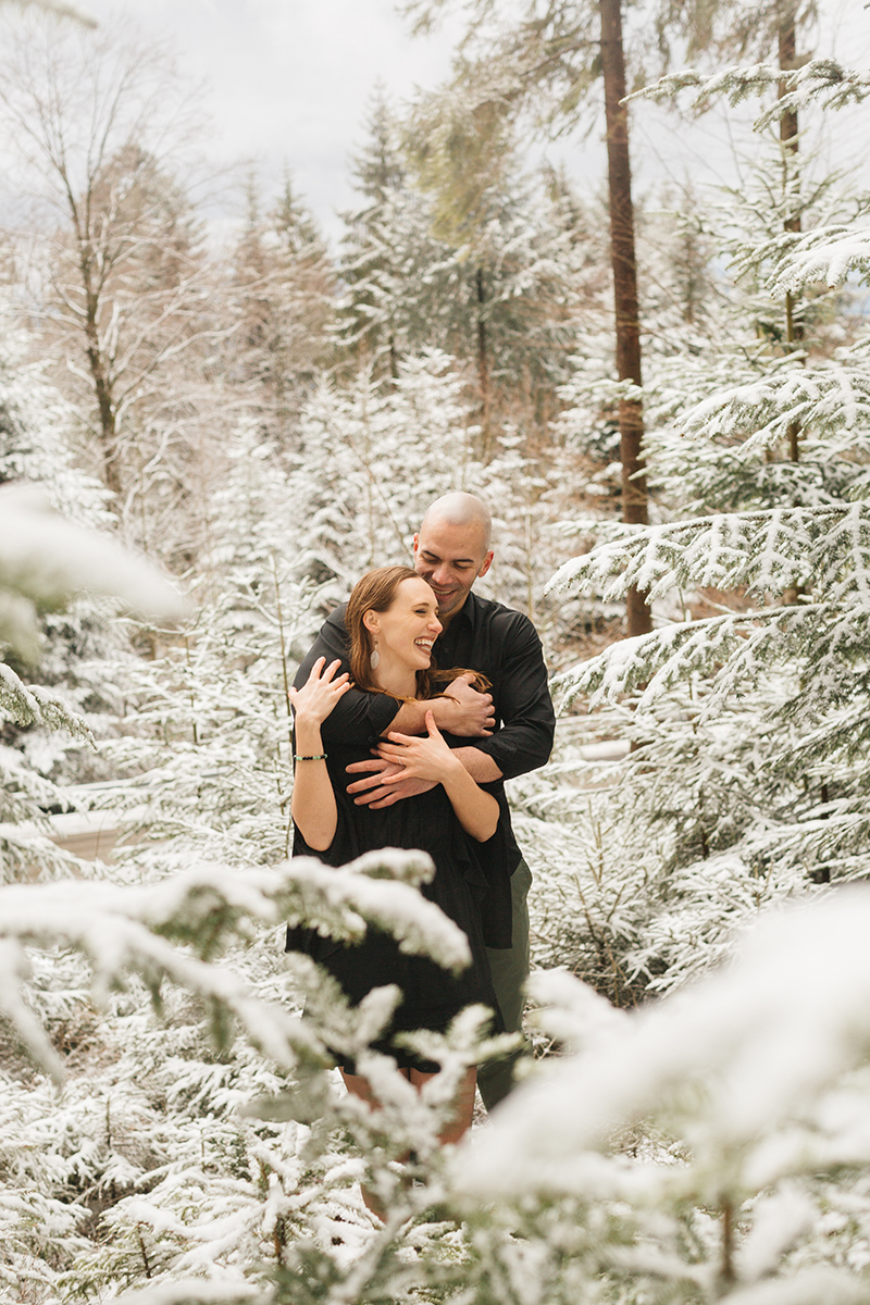 A couple embraces in a snow covered forest in Germany wearing a beautiful black dress and black button up shirt for a Black Forest engagement photography session