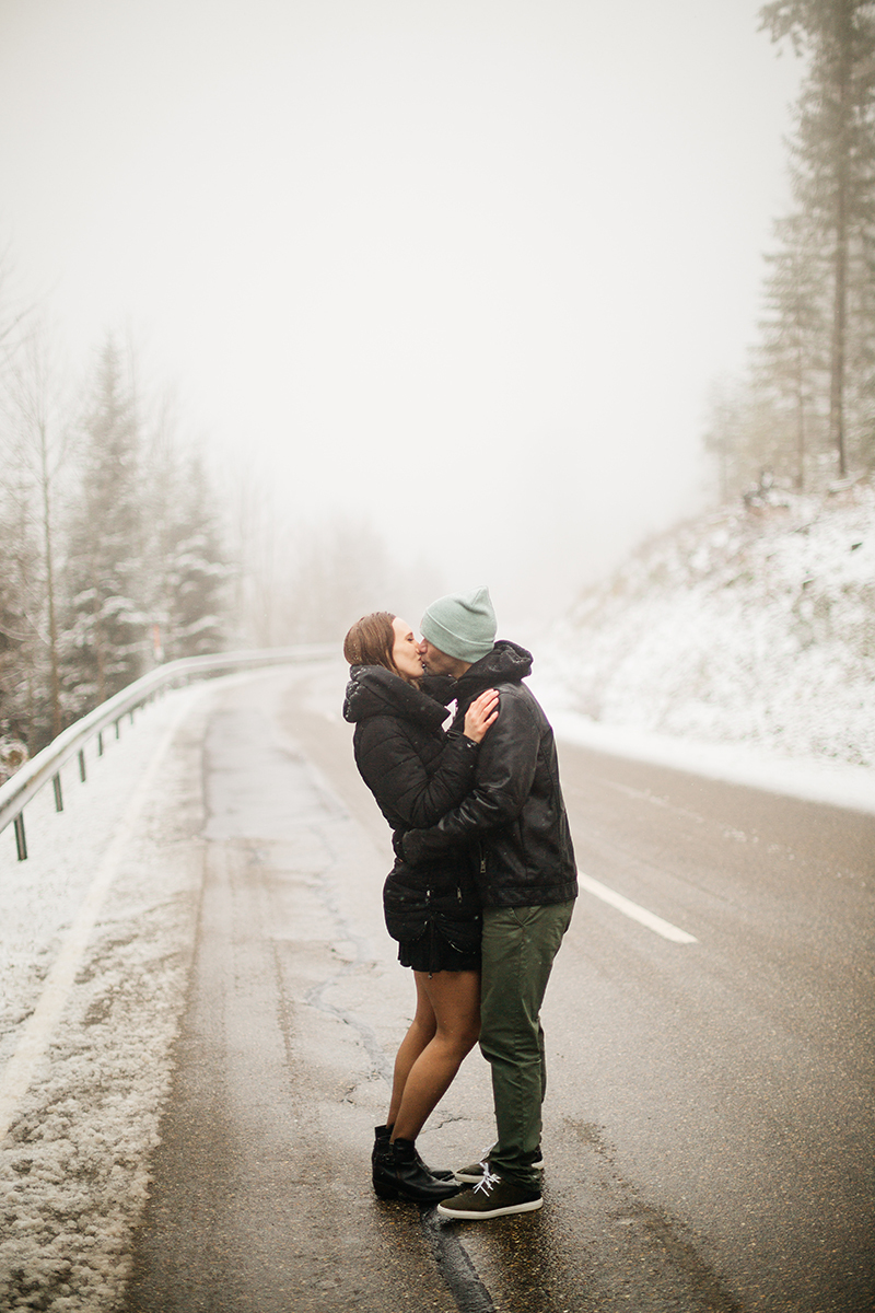 A couple embraces on a road through a snow covered forest in Germany wearing a beautiful black dress and black button up shirt for a Black Forest engagement photography session
