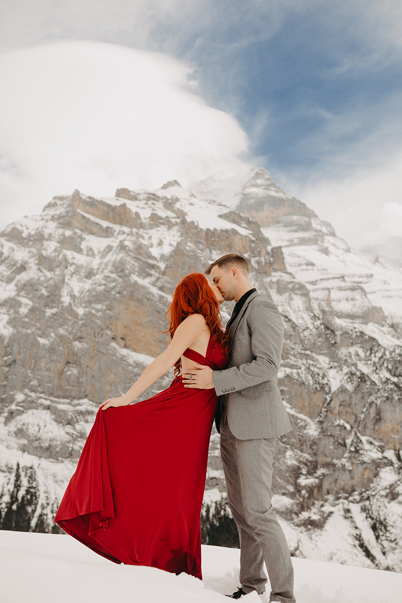 A couple kiss on a snow covered mountain in Switzerland wearing a beautiful red dress and gray suit for a Mürren couples photography session