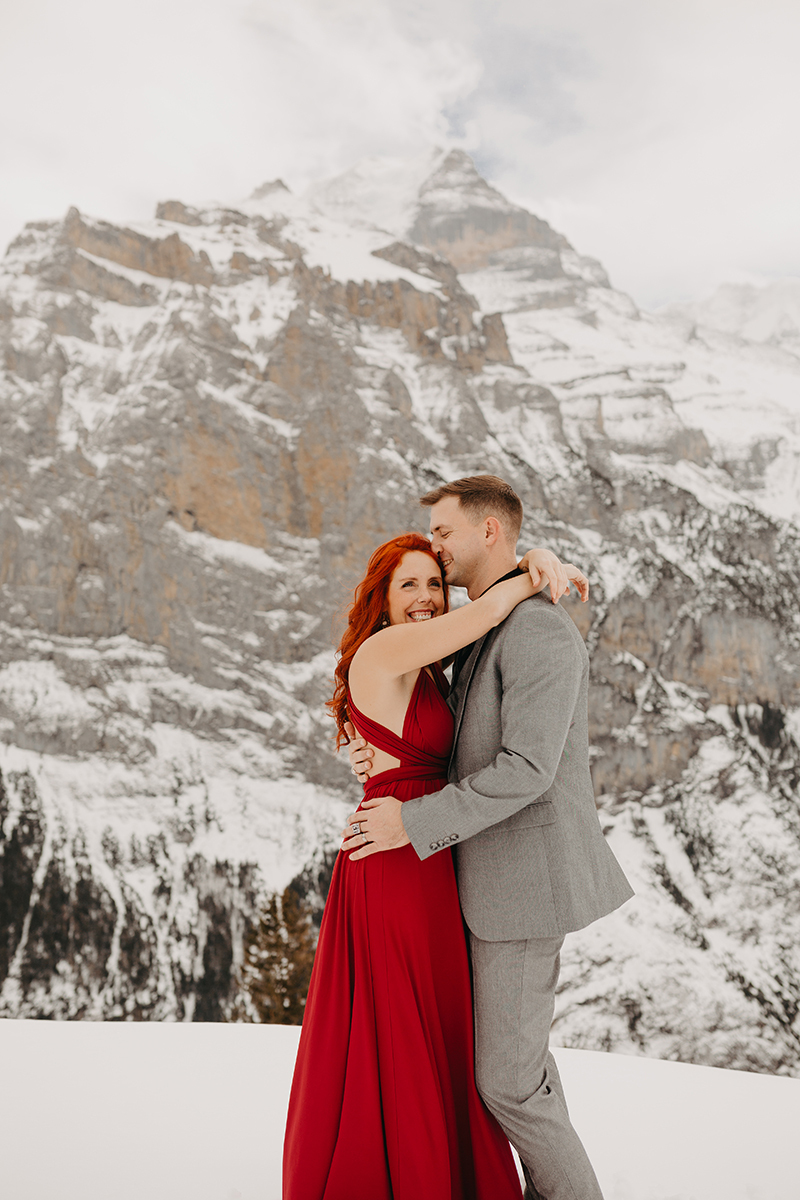 A couple embrace on a snow covered mountain in Switzerland wearing a beautiful red dress and gray suit for a Mürren couples photography session