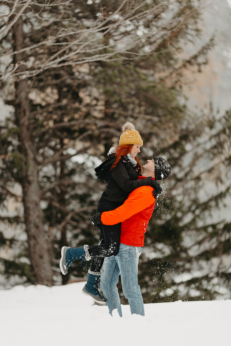 A couple embrace and kiss as he lifts her up in trees on a snow covered mountain in Switzerland wearing snow gear for a Mürren couples photography session