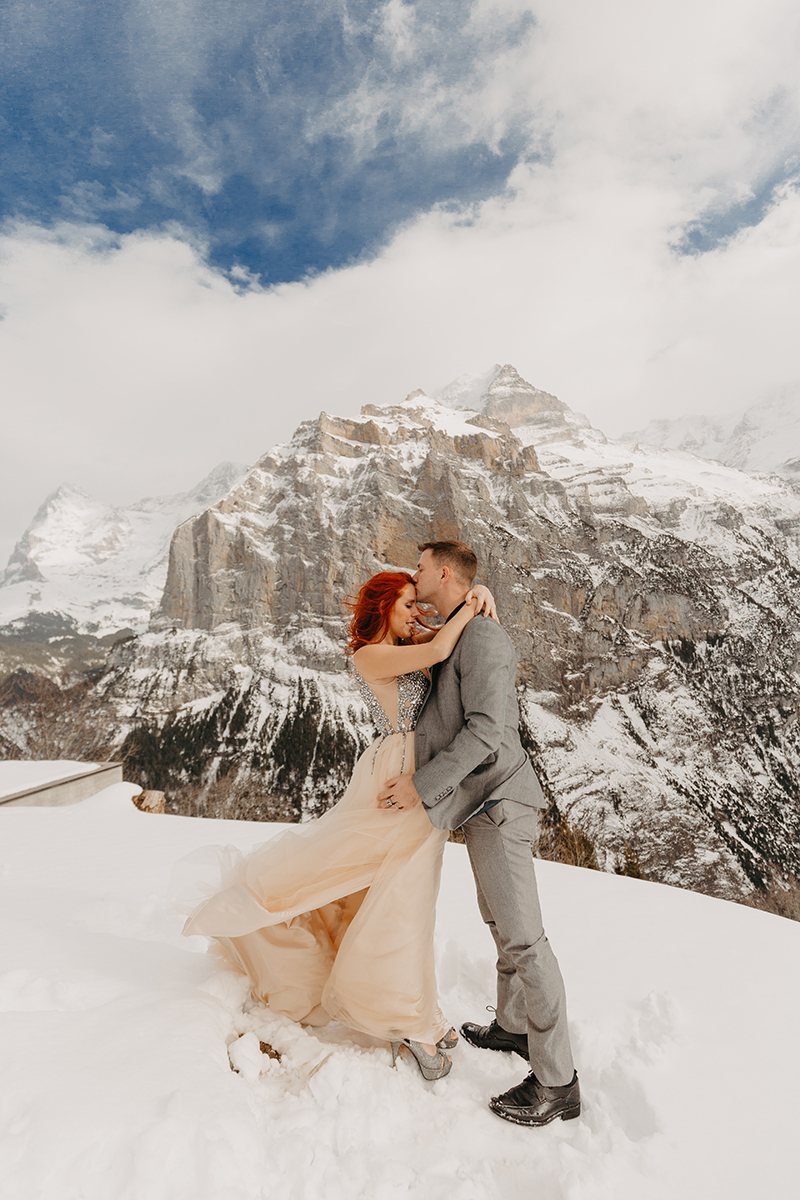 A couple embraces on a snow covered mountain in Switzerland wearing a beautiful peach colored dress and gray suit for a Mürren couples photography session
