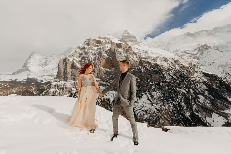 A couple stands side by side on a snow covered mountain in Switzerland wearing a beautiful peach colored dress and gray suit for a Mürren couples photography session