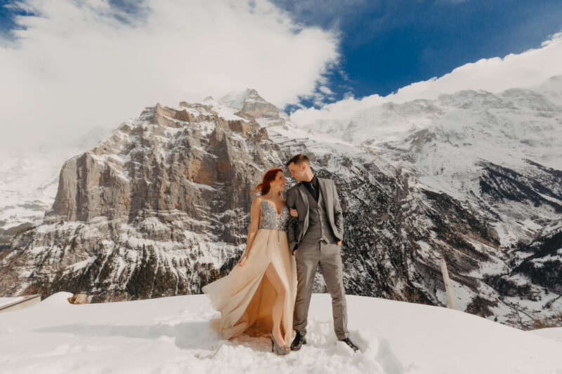 A couple stands side by side on a snow covered mountain in Switzerland wearing a beautiful peach colored dress and gray suit for a Mürren couples photography session