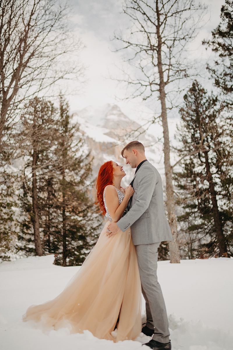 A couple embrace in trees on a snow covered mountain in Switzerland wearing a beautiful peach colored dress and gray suit for a Mürren couples photography session