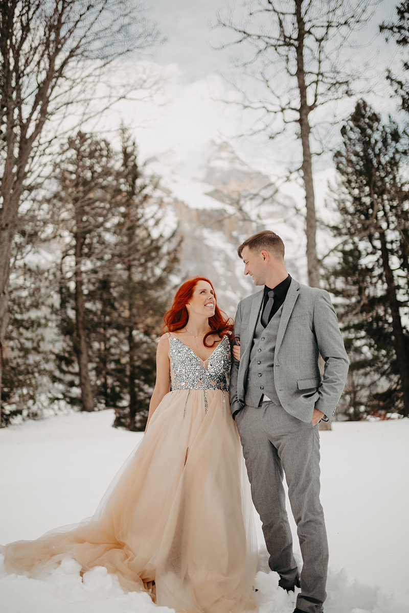 A couple walks side by side in trees on a snow covered mountain in Switzerland wearing a beautiful peach colored dress and gray suit for a Mürren couples photography session