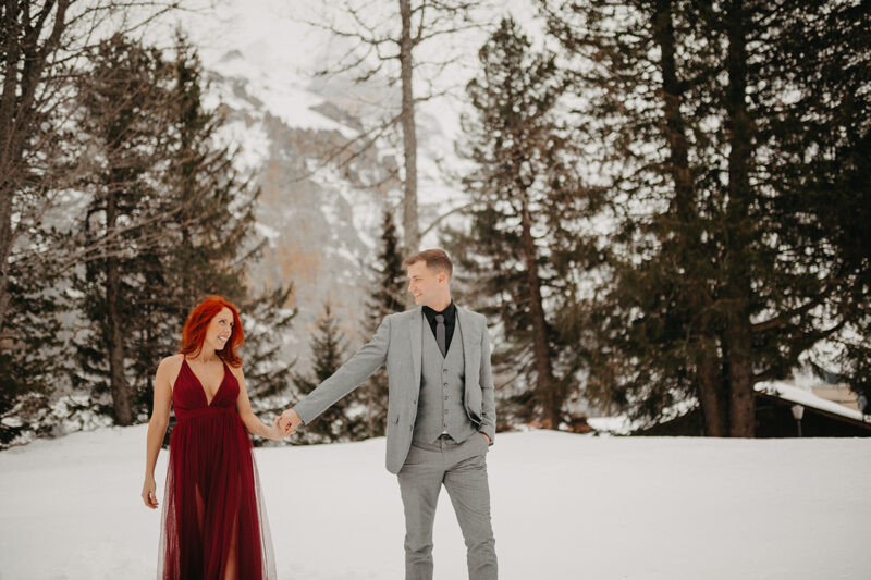 A couple walks holding hands in trees on a snow covered mountain in Switzerland wearing a beautiful red dress and gray suit for a Mürren couples photography session