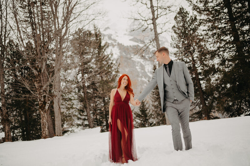 A couple walks holding hands in trees on a snow covered mountain in Switzerland wearing a beautiful red dress and gray suit for a Mürren couples photography session