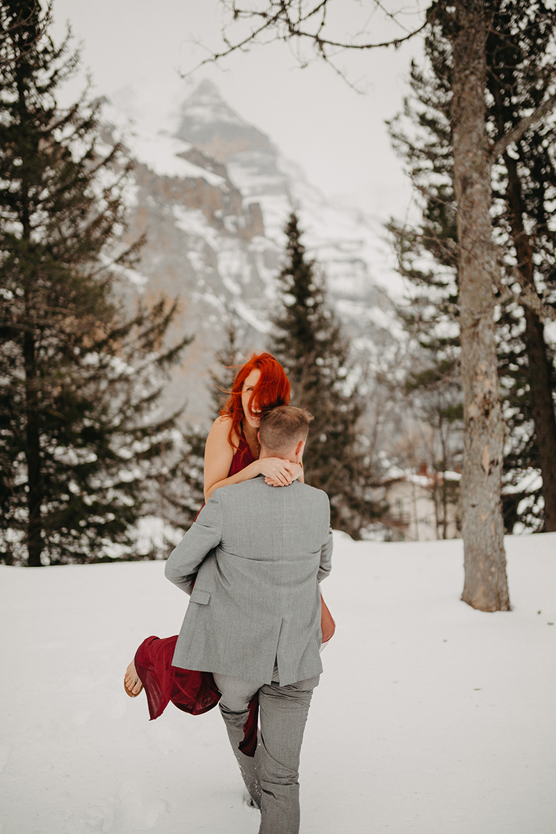 A couple walks as he carries her in trees on a snow covered mountain in Switzerland wearing a beautiful red dress and gray suit for a Mürren couples photography session