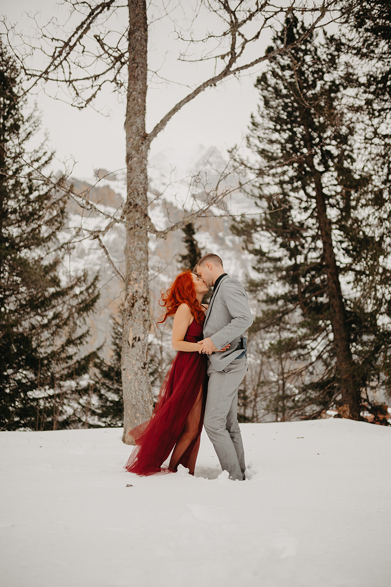 A couple embrace and kiss in trees on a snow covered mountain in Switzerland wearing a beautiful red dress and gray suit for a Mürren couples photography session