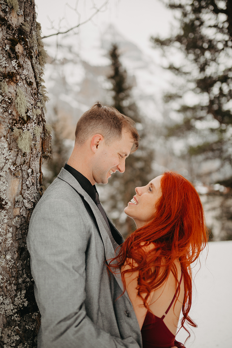 A couple embraces in trees on a snow covered mountain in Switzerland wearing a beautiful red dress and gray suit for a Mürren couples photography session